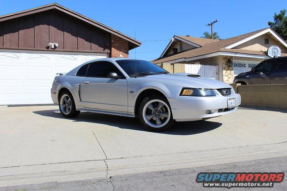 Silver Ford Mustang parked in driveway, front 3/4 view. Hood scoop and aftermarket wheels visible