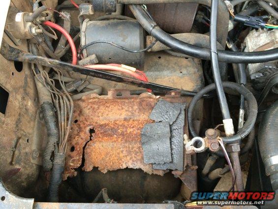 Engine bay showing a heavily rusted battery tray and wiring. Some black insulation is taped to the tray