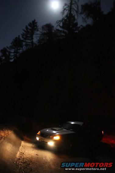 Car with headlights on, parked on a dirt road at night under a full moon