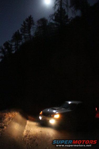 Car driving on a dirt road at night with headlights on, under a full moon