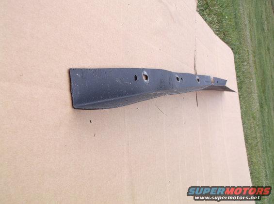 Black metal car part with holes, lying on cardboard with grass in background