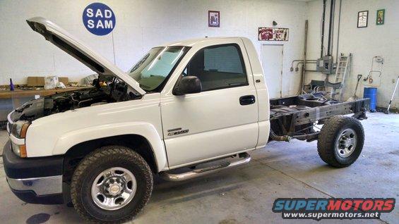 White pickup truck with hood open, showing the chassis and rear axle removed. "SUPERMOTORS.NET" logo visible