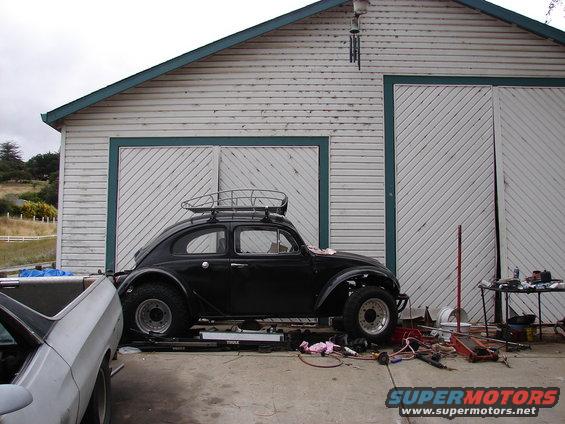 Black 1964 Volkswagen Beetle with roof rack parked in front of a garage, another car partially visible on the left