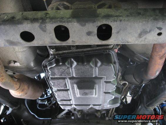 Underside view of vehicle transmission pan and exhaust pipes. Supermotors.net logo visible