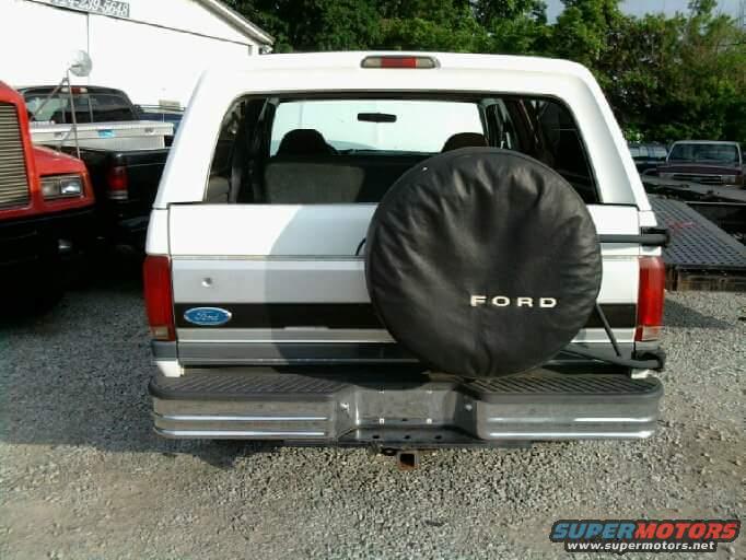 Rear view of a white 1993 Ford Bronco with a spare tire on the back. A red truck is to the left