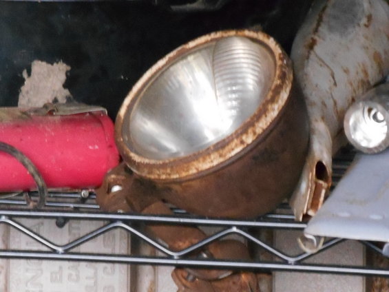 Rusty headlight assembly with ribbed glass lens, next to a red cylindrical object