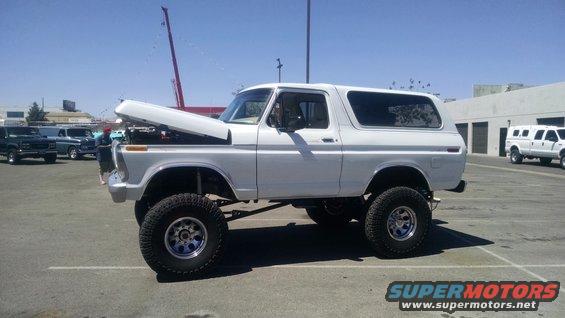 Lifted white 1978 Ford Bronco parked outside with hood open. Other vehicles and buildings in background