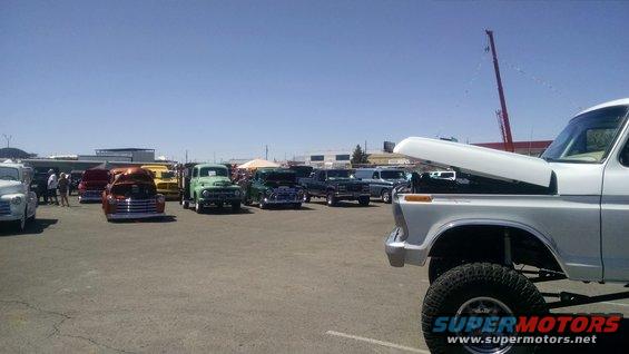Several classic trucks parked in a lot under a clear sky. A white pickup with its hood up is in the foreground