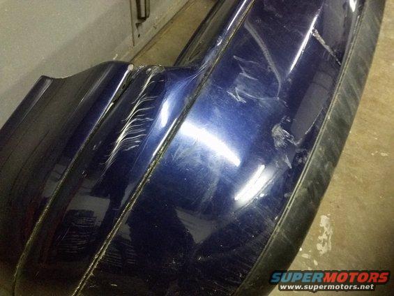 Damaged blue bumper with deep scratches and dents, showing a black trim piece