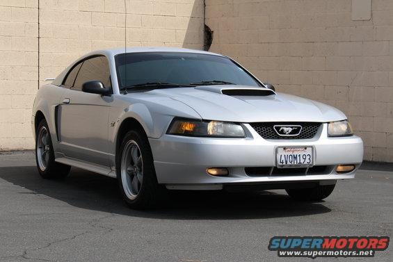 Silver 2002 Ford Mustang coupe with a hood scoop and GT badging, parked in front of a beige brick wall