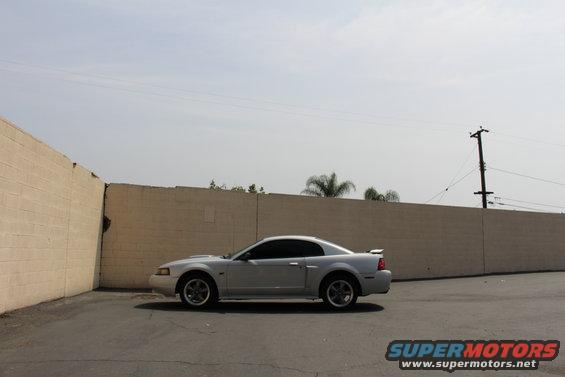 Silver 2002 Ford Mustang coupe parked in a lot next to a tan brick wall
