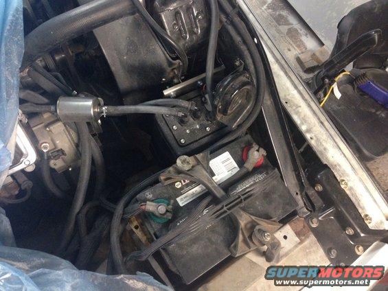 Car battery, fuel filter, and some hoses in an engine bay. Some blue plastic is covering parts