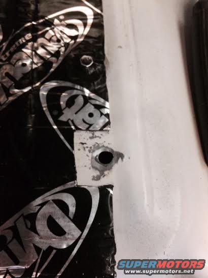 Black sound deadening material with silver logos is adhered to a white car door panel with a drilled hole