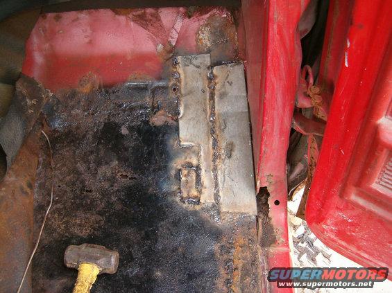 Red vehicle interior floor pan with extensive rust and welded metal patches. A sledgehammer rests on the floor