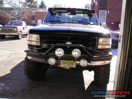 Front view of black Ford F-Series truck with a brush guard and four lights. Another red truck is parked behind it