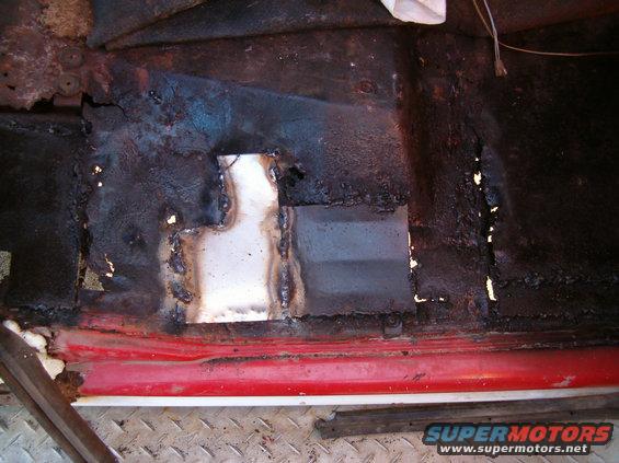 Rusted floor pan with a welded patch panel, red trim visible below