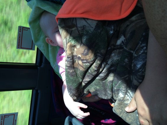 Baby sleeping, covered by a camouflage blanket, with an orange shirt visible. Two 'EMERGENCY EXIT' stickers are on the window
