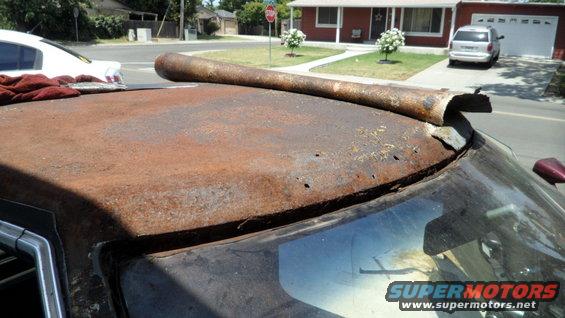 Rusted car roof with holes and a bent metal pipe on top, showing a white car and a minivan in the background