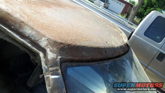 Rusted roofline and windshield frame of a vehicle, with a pickup truck visible behind