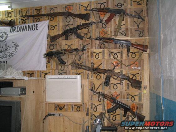 Wall display of several rifles and machine guns mounted on wooden crates. A white flag with the word "ORDNANCE" is on the left