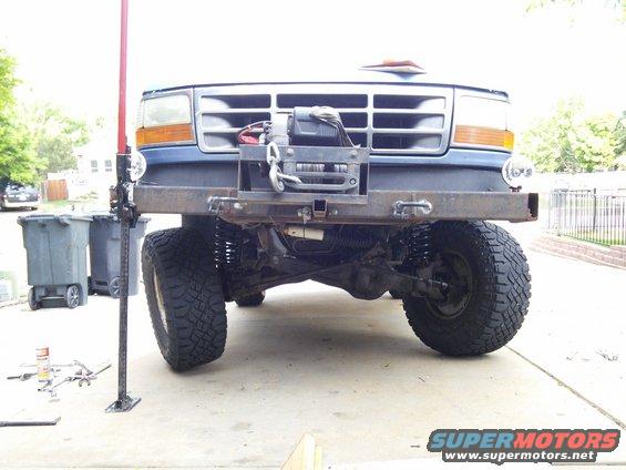 Front of a blue Ford F-Series truck with a winch mounted on a custom bumper. A Hi-Lift jack is positioned next to it