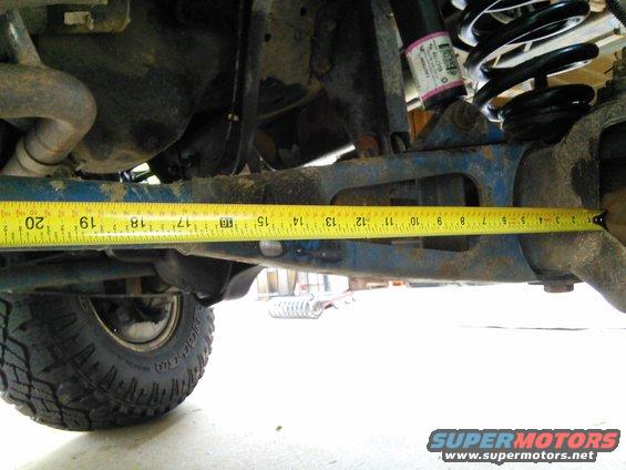 Measuring tape across blue suspension arm on vehicle underside. Coil spring and tire visible
