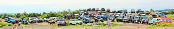 Many off-road vehicles parked on a rocky, grassy hill under a cloudy sky