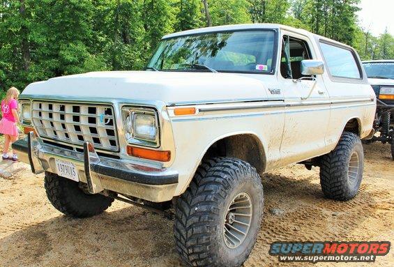 Lifted white Ford Bronco with big tires, a girl in a pink skirt stands nearby. Another vehicle is partially visible behind it
