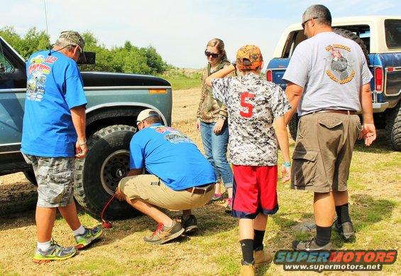 People gathered around a 1979 Ford Bronco, one person is airing up the tire. Another Bronco is parked behind it