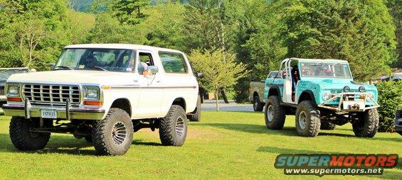 White Bronco and a teal Bronco parked on grass with trees in background. White Bronco has a license plate that reads 1979015
