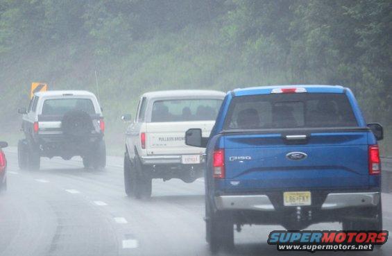Rear view of a white Ford Bronco with a spare tire on the back, driving in the rain