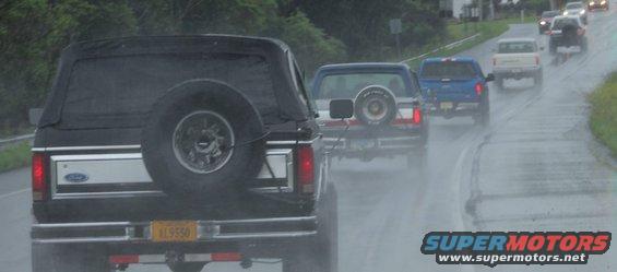 Rear view of a black Ford Bronco with a spare tire on the back, driving in the rain behind other vehicles