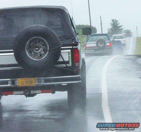 Rear view of a black Ford Bronco with a spare tire on the back, driving on a wet road behind other vehicles