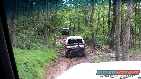 White 1979 Ford Bronco driving on a rocky forest trail, followed by another SUV