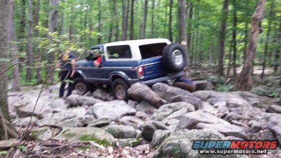 Blue SUV with white top driving over large rocks in a forest