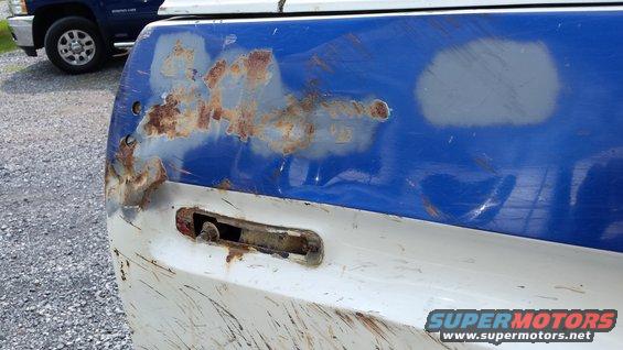 rusted blue and white tailgate corner, missing taillight assembly. Blue pickup truck in background