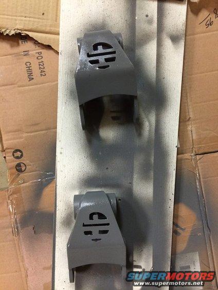 Two gray painted suspension parts with a circular logo, resting on a wooden board over cardboard