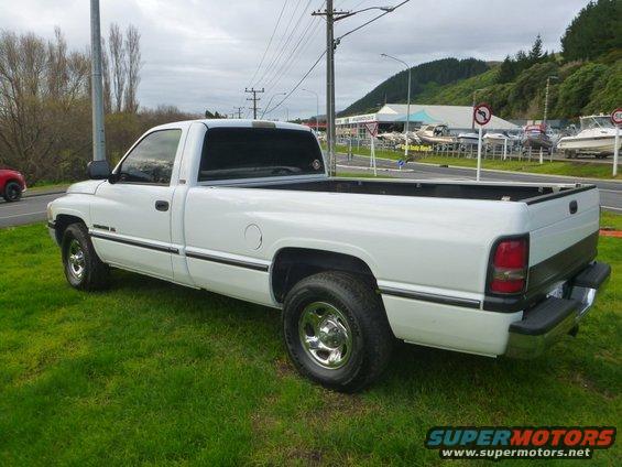 White pickup truck parked on grass, side view. Background shows a road, buildings, and boats