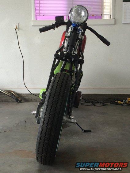 Motorcycle front end with large headlight, black handlebars, and exposed front suspension. A red fuel tank is partially visible
