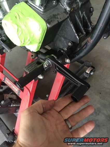 Hand holding a black motorcycle kickstand lever near a black engine block covered in green tape, on a red Craftsman lift
