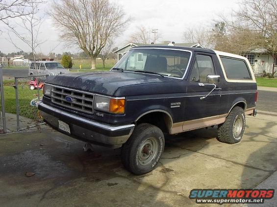 Dark blue Ford Bronco with tan trim, parked on a driveway. Another SUV is visible in the background