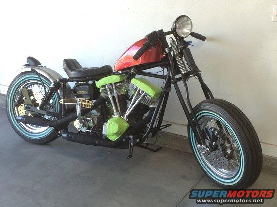 Chopper motorcycle with red fuel tank, black seat, and green engine covers. Whitewall tires and chrome exhaust are visible