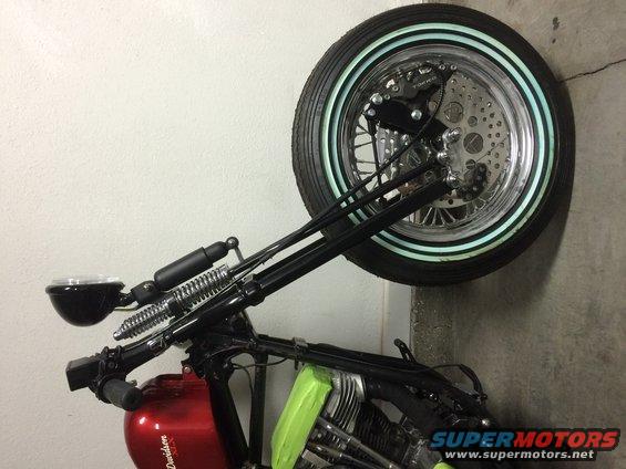 Motorcycle front end with black handlebars, dual springs, and a chrome wheel with a whitewall tire. Red fuel tank visible
