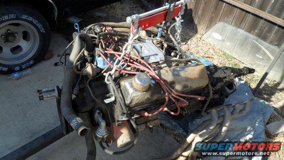 Engine assembly with red engine hoist chains, chrome valve cover, and spark plug wires. A tire and windshield are visible nearby