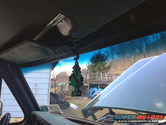 Interior view from driver's seat looking out windshield, showing a Black Forest air freshener and the open hood of a light-colored vehicle