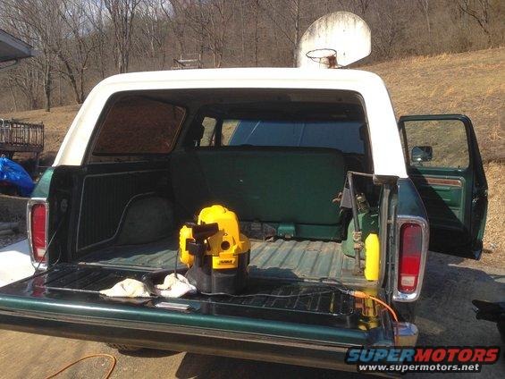 Rear interior of a 1978 Ford Bronco with a yellow shop vac and cleaning supplies. The tailgate is down and the driver's door is open