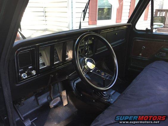 Interior shot of a truck's dashboard and steering wheel. The dashboard has rectangular gauges and a radio. The steering wheel is black with chrome accents