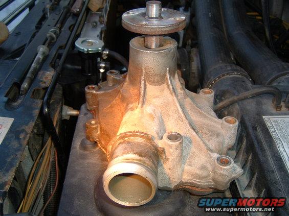 Close-up of a water pump assembly with a shaft protruding from the top, situated within an engine bay