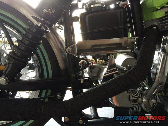 Close-up of a motorcycle's black exhaust pipe, rear shock, and engine components. The fender has green stripes