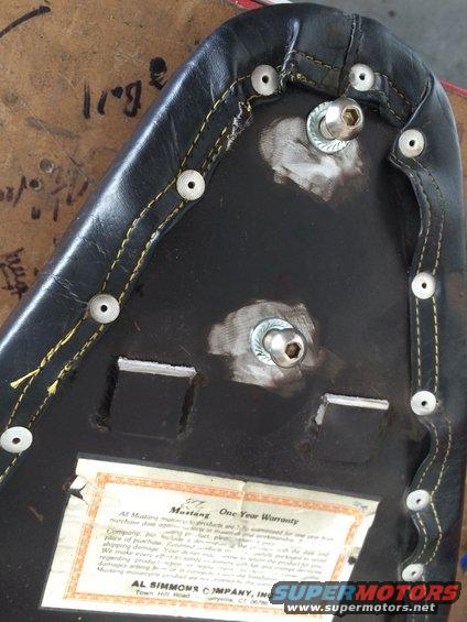 Underside of a black motorcycle seat with yellow stitching and metal fasteners. A warranty tag is visible